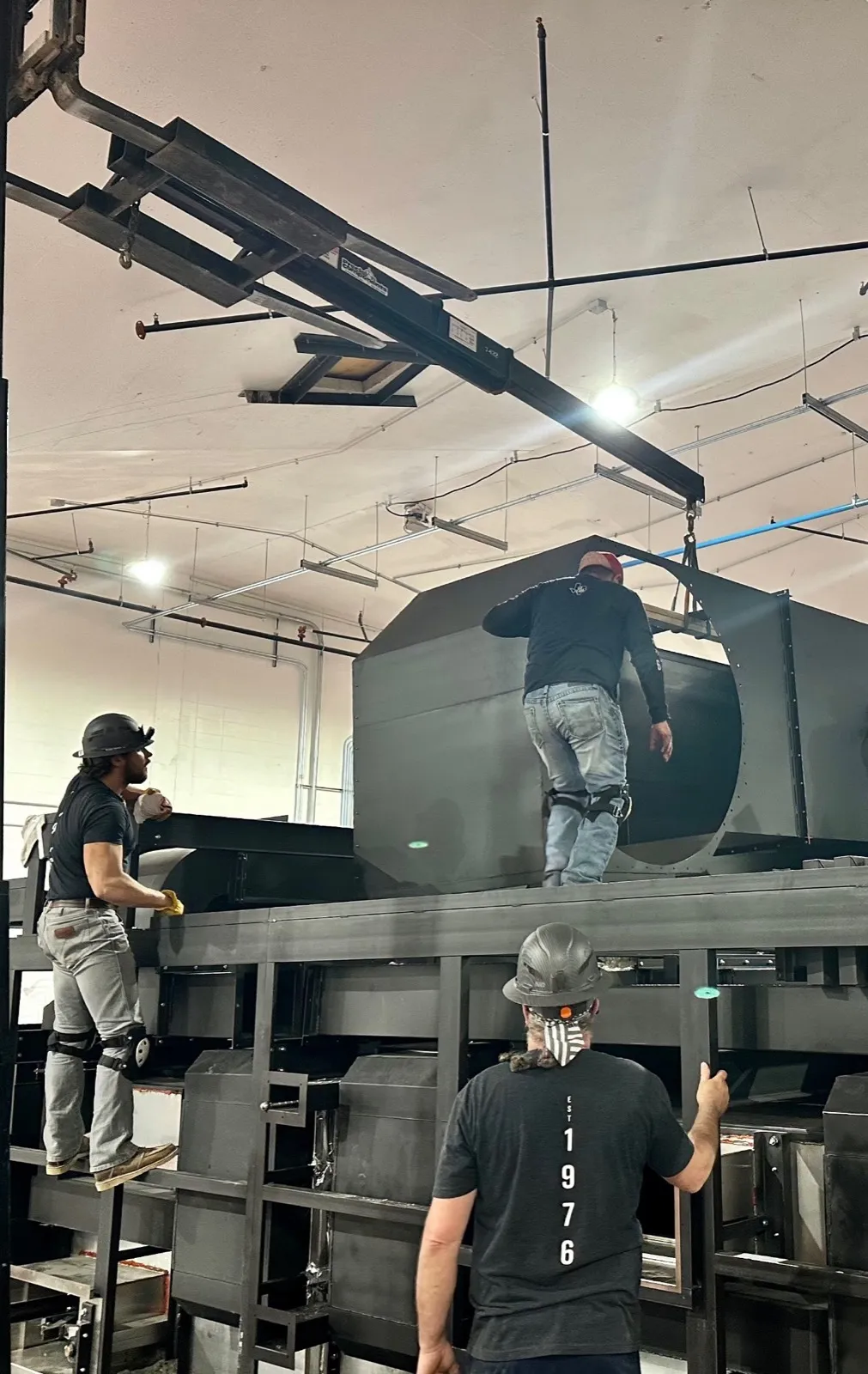 Three Henry Group crew members assemble a large oven module, one working on top of the frame and another steadying it from a ladder.