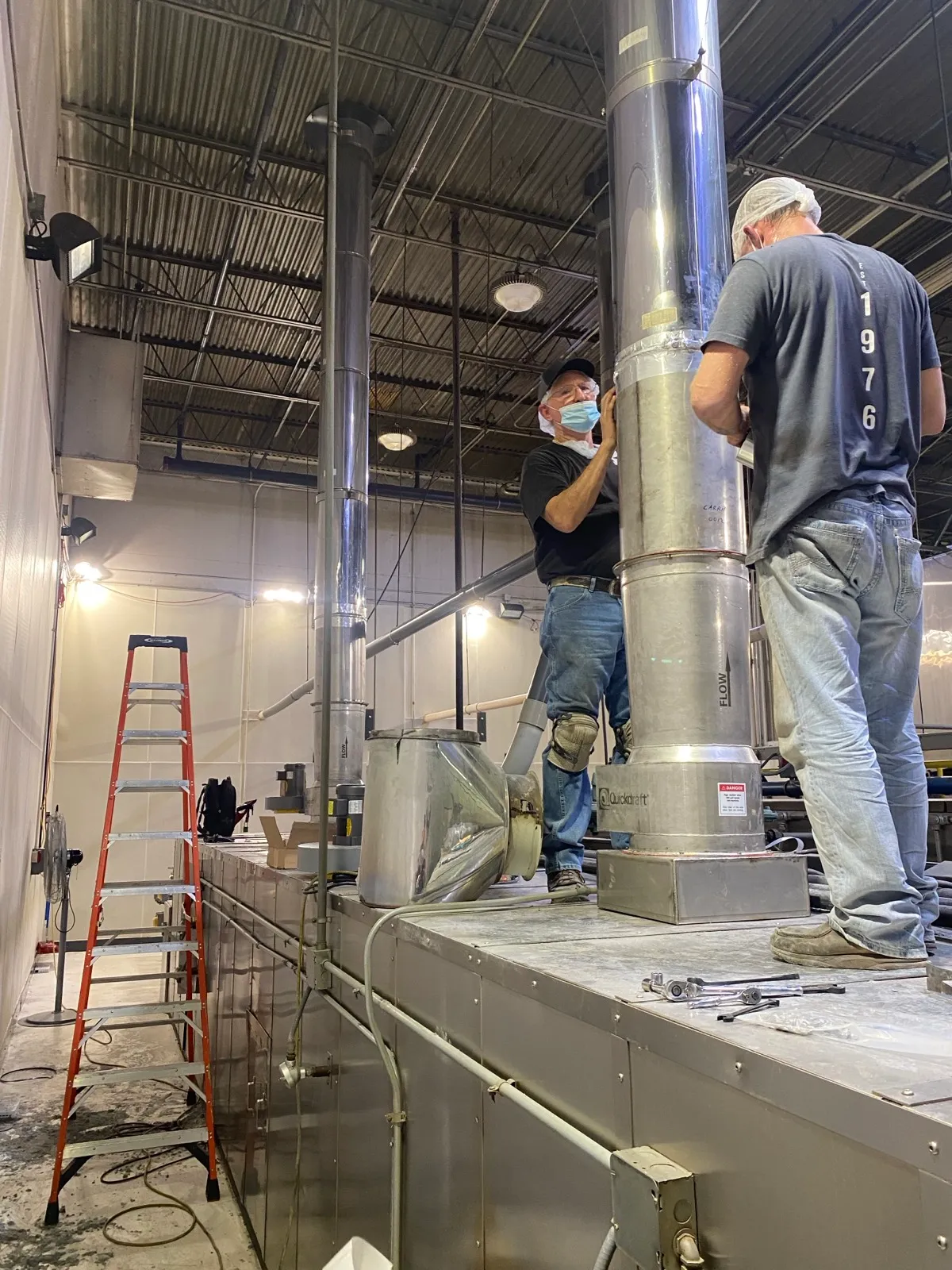 Two Henry Group technicians fit a stainless exhaust stack to the top of a bakery oven, with a ladder and hand tools staged alongside.