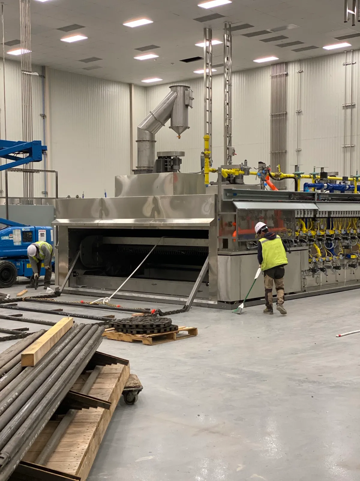 Wide view of an oven installation in progress, with two Henry Group workers, a scissor lift, and conveyor belting and chain staged across the plant floor.