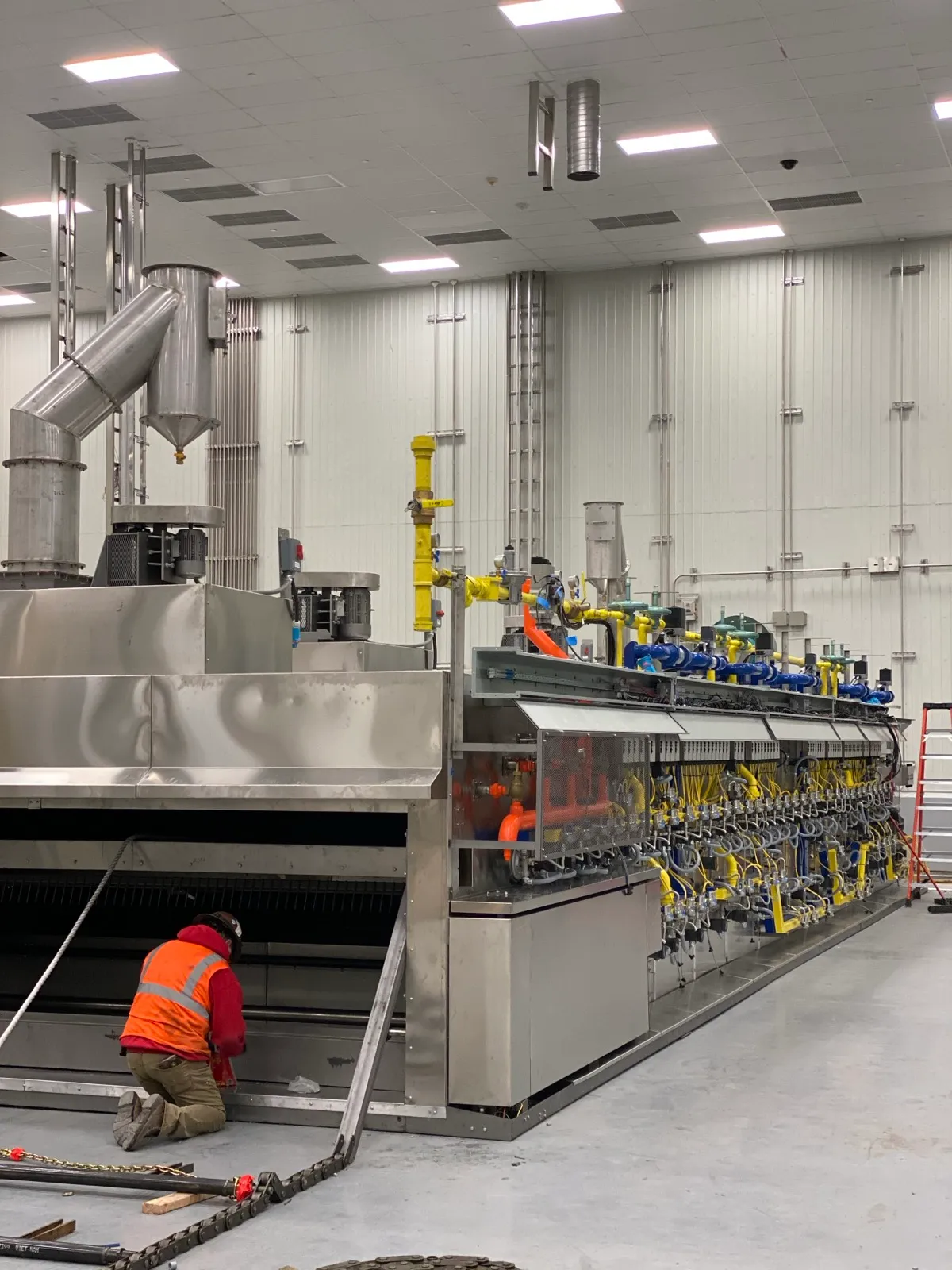 A Henry Group technician in a hi-vis vest kneels at the infeed of a newly installed industrial tunnel oven, the full burner manifold running the length of the line behind him.