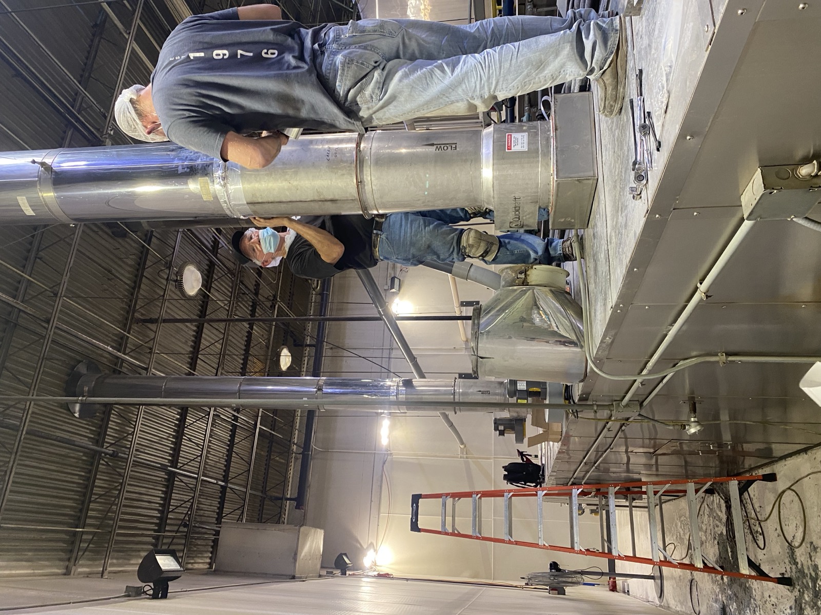 Two Henry Group technicians fit a stainless exhaust stack to the top of a bakery oven, with a ladder and hand tools staged alongside.