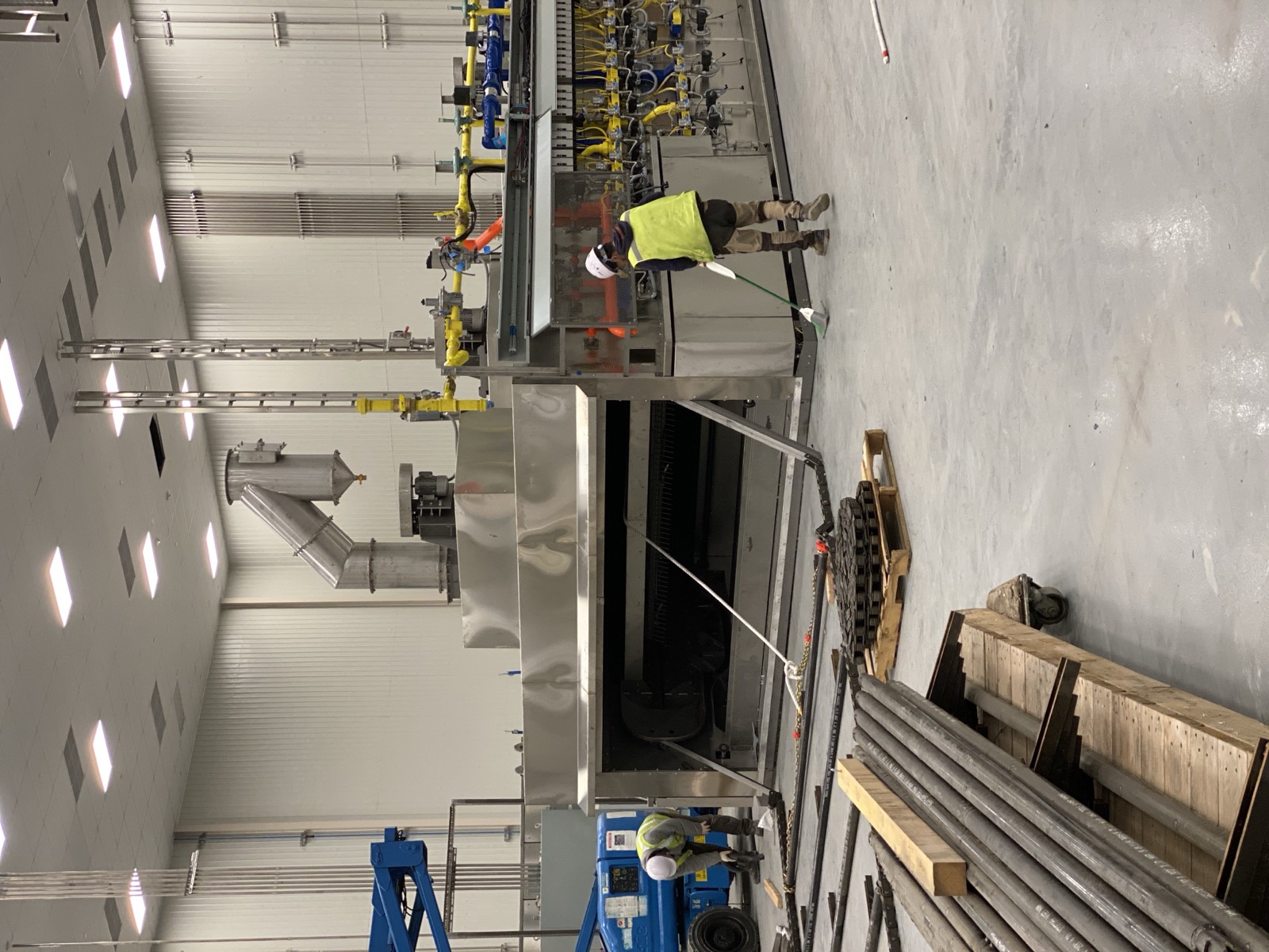 Wide view of an oven installation in progress, with two Henry Group workers, a scissor lift, and conveyor belting and chain staged across the plant floor.