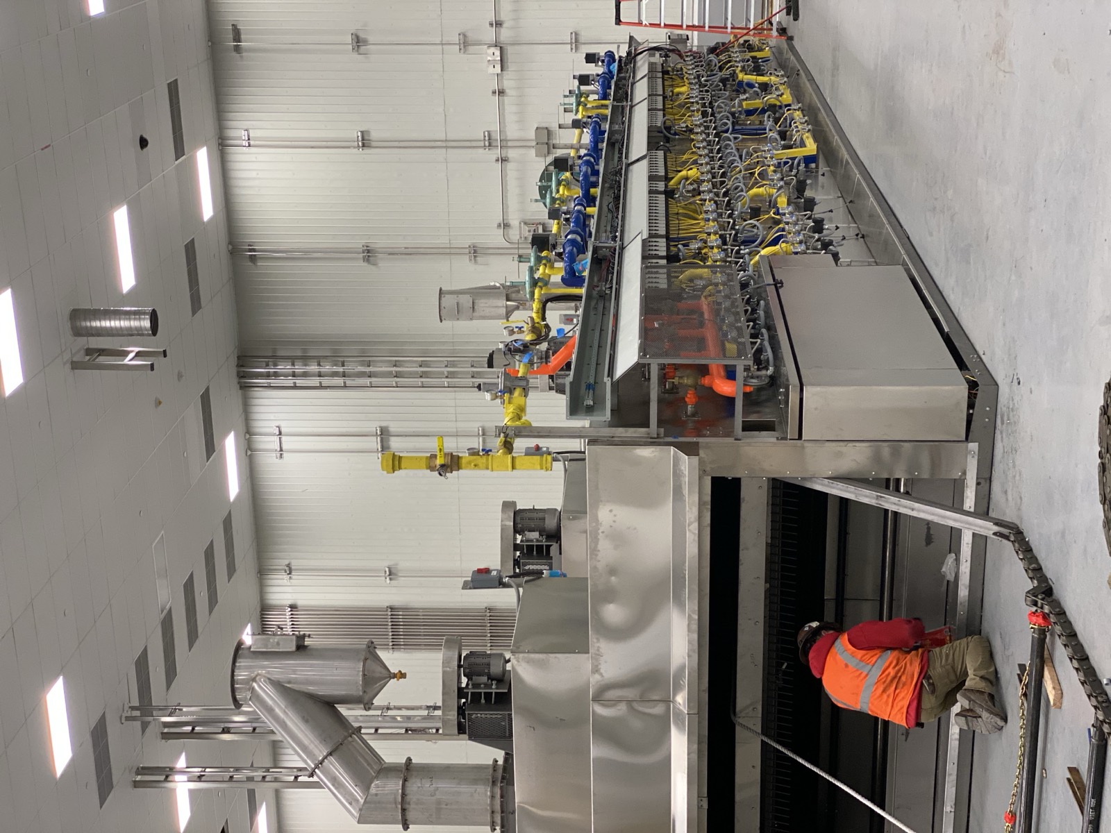 A Henry Group technician in a hi-vis vest kneels at the infeed of a newly installed industrial tunnel oven, the full burner manifold running the length of the line behind him.